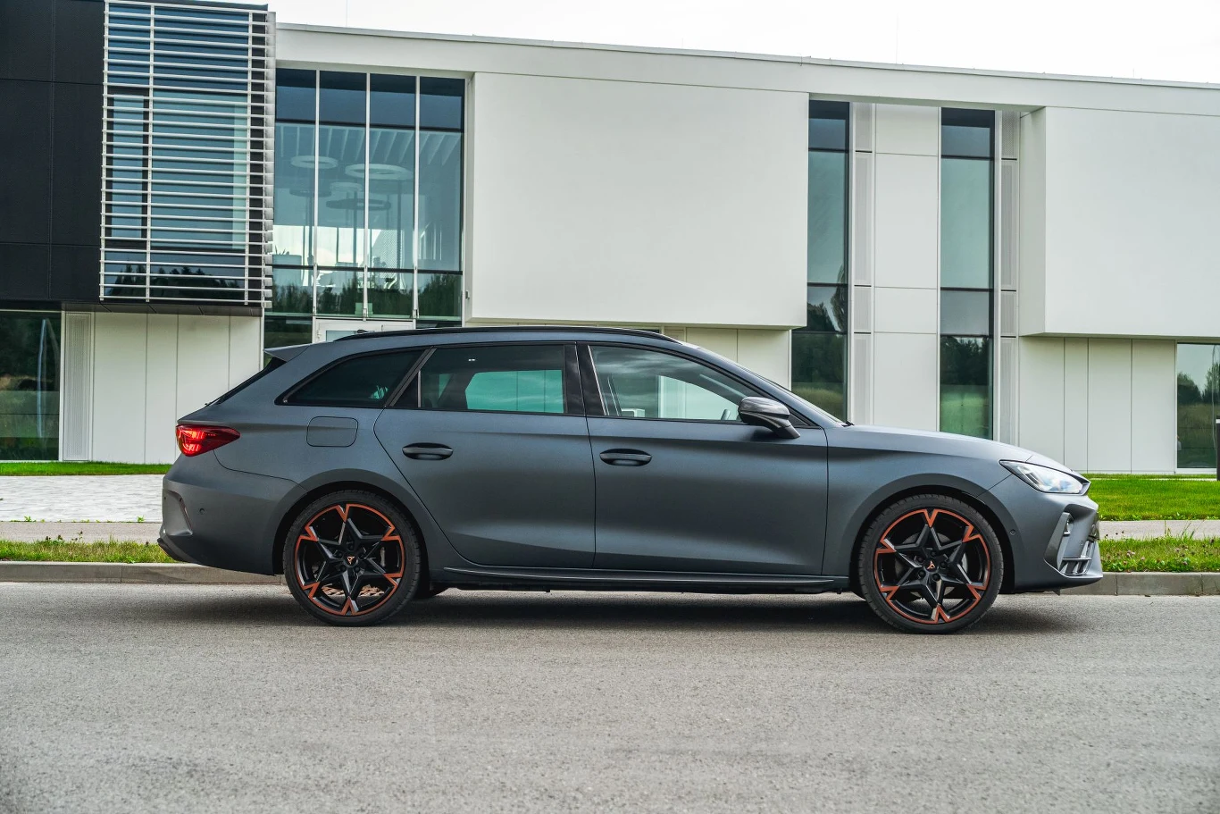 Po modernizacji średnie deklarowane przez producenta zużycie paliwa Cupry Leon z 204-konną hybrydą wynosi 0,4 l/100 km. Nowoczesny samochód osobowy w kolorze matowej szarości zaparkowany na ulicy przed białym, przeszklonym budynkiem, wyróżniający się czarnymi felgami z czerwonymi akcentami.