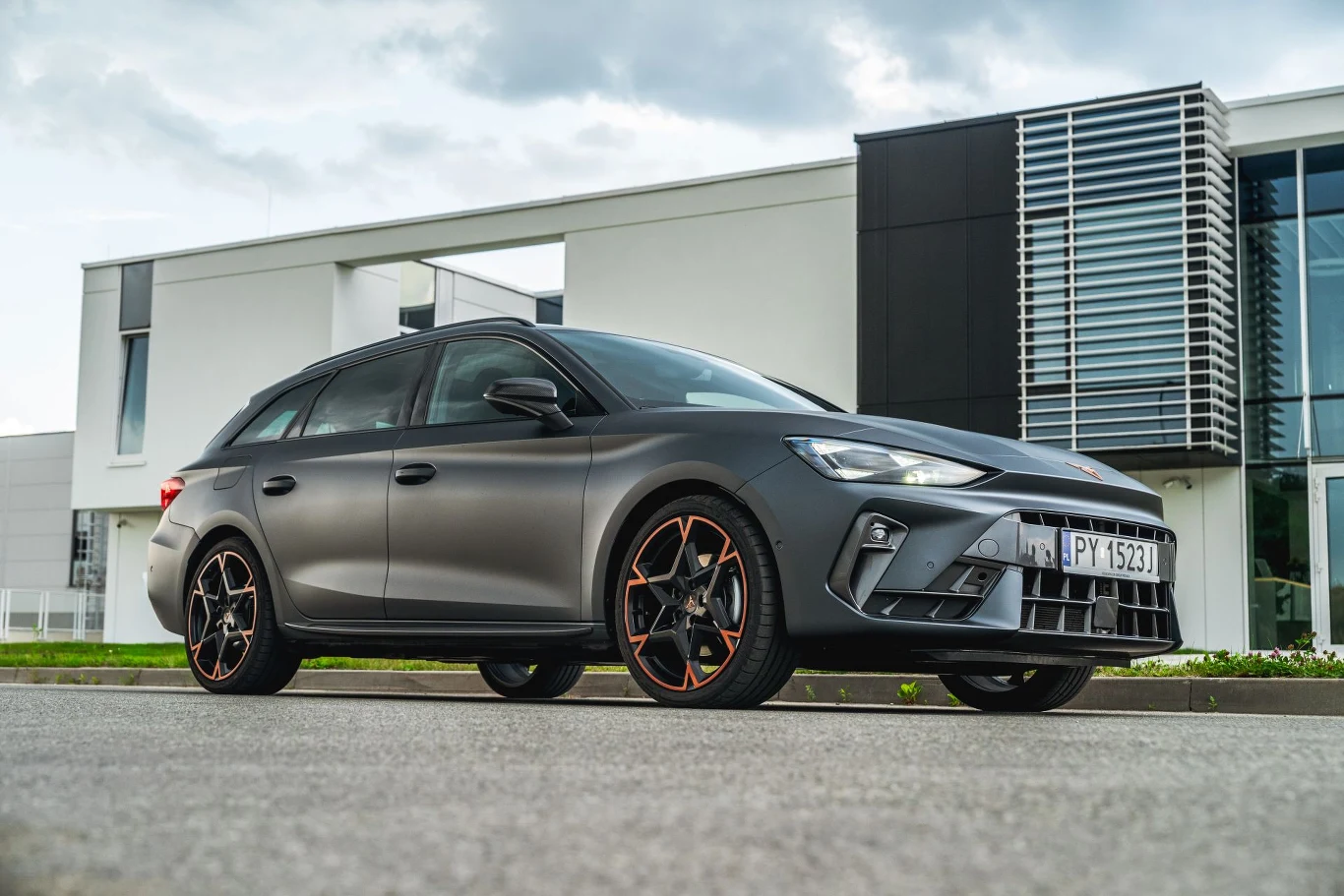 Umieszczony w tylnej części auta akumulator zapewnia stabilność przy pokonywaniu zakrętów. Nowoczesny, sportowy samochód osobowy o matowym, szarym lakierze stoi zaparkowany na tle nowoczesnego budynku z dużymi przeszkleniami i geometrycznymi elewacjami.