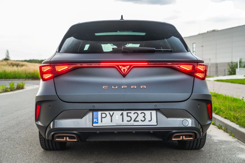 Cupra Leon z 204-konną hybrydą plug-in dostępna jest tylko w standardowej wersji wyposażenia. Nowoczesny samochód marki Cupra widziany od tyłu, ze światłami tylnymi LED, tablicą rejestracyjną PY 1523J i sportowym dyfuzorem wydechu, ustawiony na asfaltowej, pustej drodze obok niskich traw i nowoczesnego budynku.