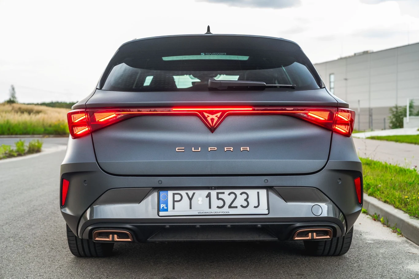 Cupra Leon z 204-konną hybrydą plug-in dostępna jest tylko w standardowej wersji wyposażenia. Nowoczesny samochód marki Cupra widziany od tyłu, ze światłami tylnymi LED, tablicą rejestracyjną PY 1523J i sportowym dyfuzorem wydechu, ustawiony na asfaltowej, pustej drodze obok niskich traw i nowoczesnego budynku.