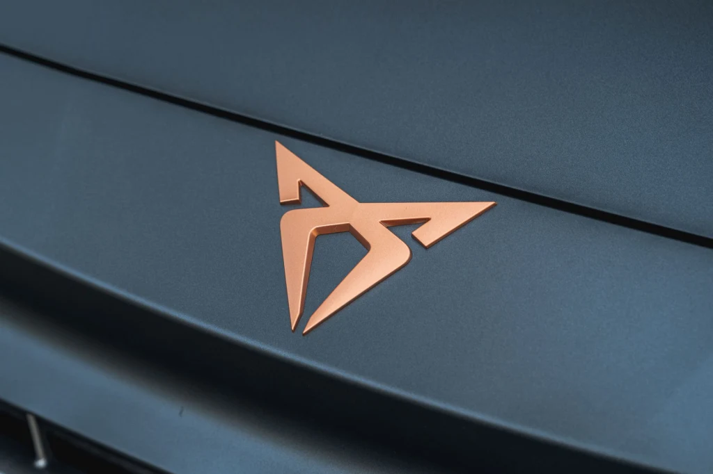 Cupra Leon z 204-konną hybrydą plug-in kosztuje 178 700 zł. Logo marki Cupra w kolorze miedzianym na matowej, ciemnej powierzchni maski samochodu.