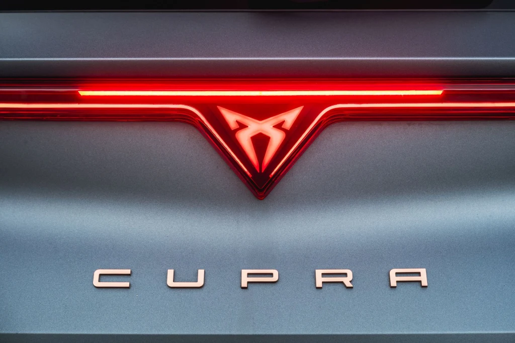 Tylne światła Cupry Leon wciąż połączone są świetlną listwą, ale teraz na jej środku znajduje się podświetlane logo. Logo Cupra podświetlone na czerwono i napis CUPRA na tylnej części samochodu na tle metalicznej karoserii.