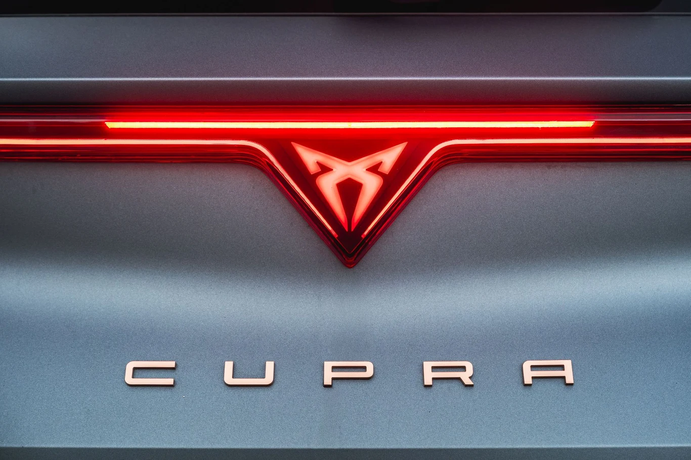 Tylne światła Cupry Leon wciąż połączone są świetlną listwą, ale teraz na jej środku znajduje się podświetlane logo. Logo Cupra podświetlone na czerwono i napis CUPRA na tylnej części samochodu na tle metalicznej karoserii.