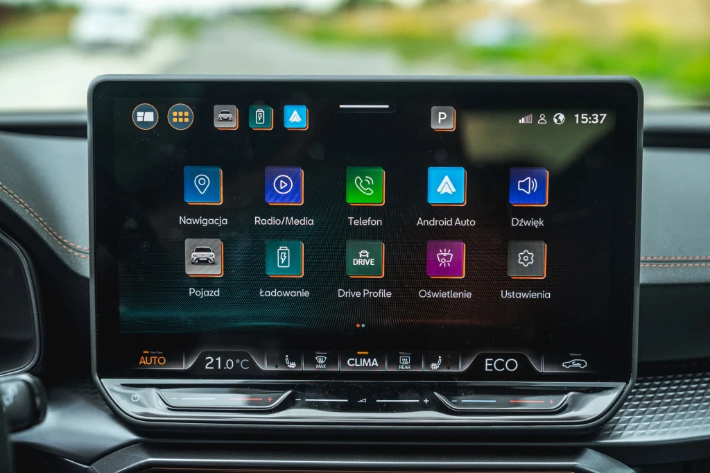 Nowy system multimediów obsługujemy z poziomu 12,9-calowego ekranu. Nowoczesny ekran systemu multimedialnego samochodu z wyraźnie widocznymi ikonami aplikacji do nawigacji, radia, telefonu, Android Auto, ustawień oraz klimatyzacji. Panel sterowania klimatyzacją i ekologicznym trybem jazdy znajdują się pod ekranem dotyk...