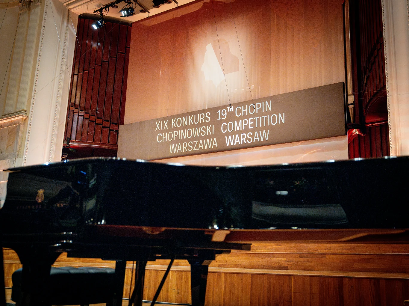 Ile kosztuje fortepian Steinway & Sons na Konkursie Chopinowskim? Sala koncertowa z fortepianem ustawionym na scenie, w tle widoczna duża tablica informacyjna z napisem XIX Konkurs Chopinowski Chopin Competition Warsaw Warszawa podświetlona reflektorami.
