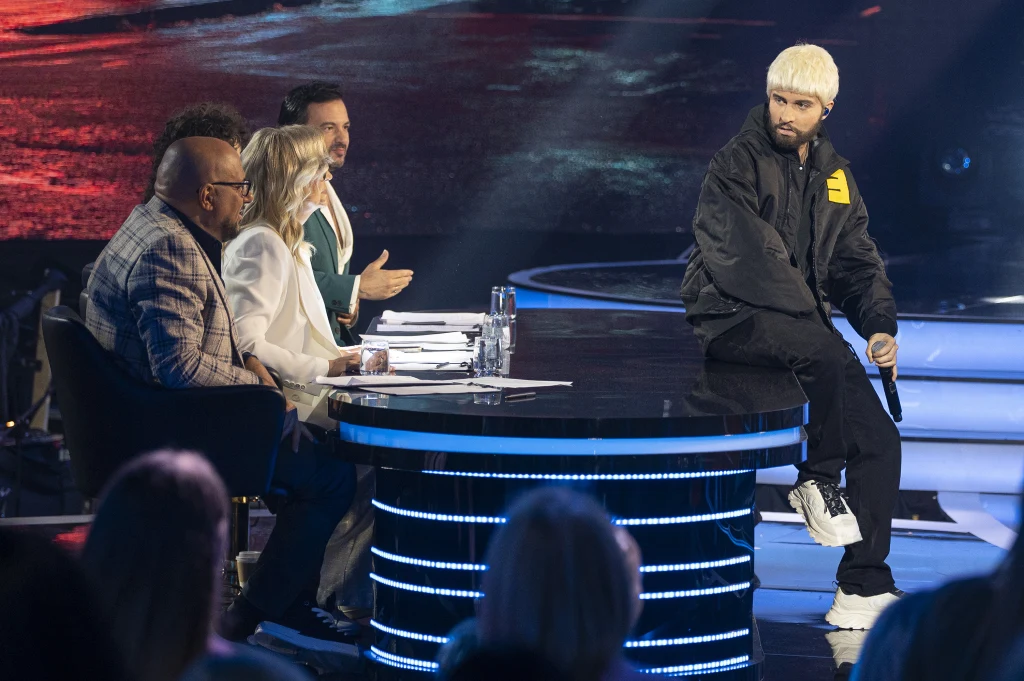 Kamil Studnicki jako Eminem w programie "Twoja twarz brzmi znajomo" Czworo jurorów siedzących przy okrągłym stole podczas programu rozrywkowego w jasnym oświetleniu sceny, jeden uczestnik w jasnej fryzurze stoi bokiem przy stole zwracając na siebie uwagę.