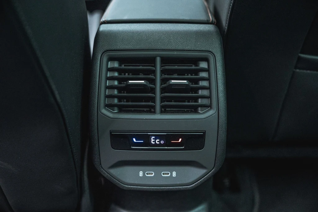 Już w standardowej wersji wyposażenia Cupra Leon jest wyposażona w trzystrefową klimatyzację automatyczną. Panel tylnej klimatyzacji samochodu z widocznymi kratkami wentylacyjnymi oraz cyfrowym wskaźnikiem trybu Eco i regulacją temperatury, otoczony czarnym wnętrzem pojazdu.