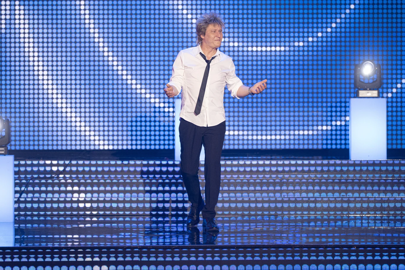 Mężczyzna występujący na scenie oświetlonej niebieskimi światłami LED, ubrany w białą koszulę z rozpiętym krawatem, czarne spodnie i czarne buty, w dynamicznej pozie sugerującej ruch lub taniec.