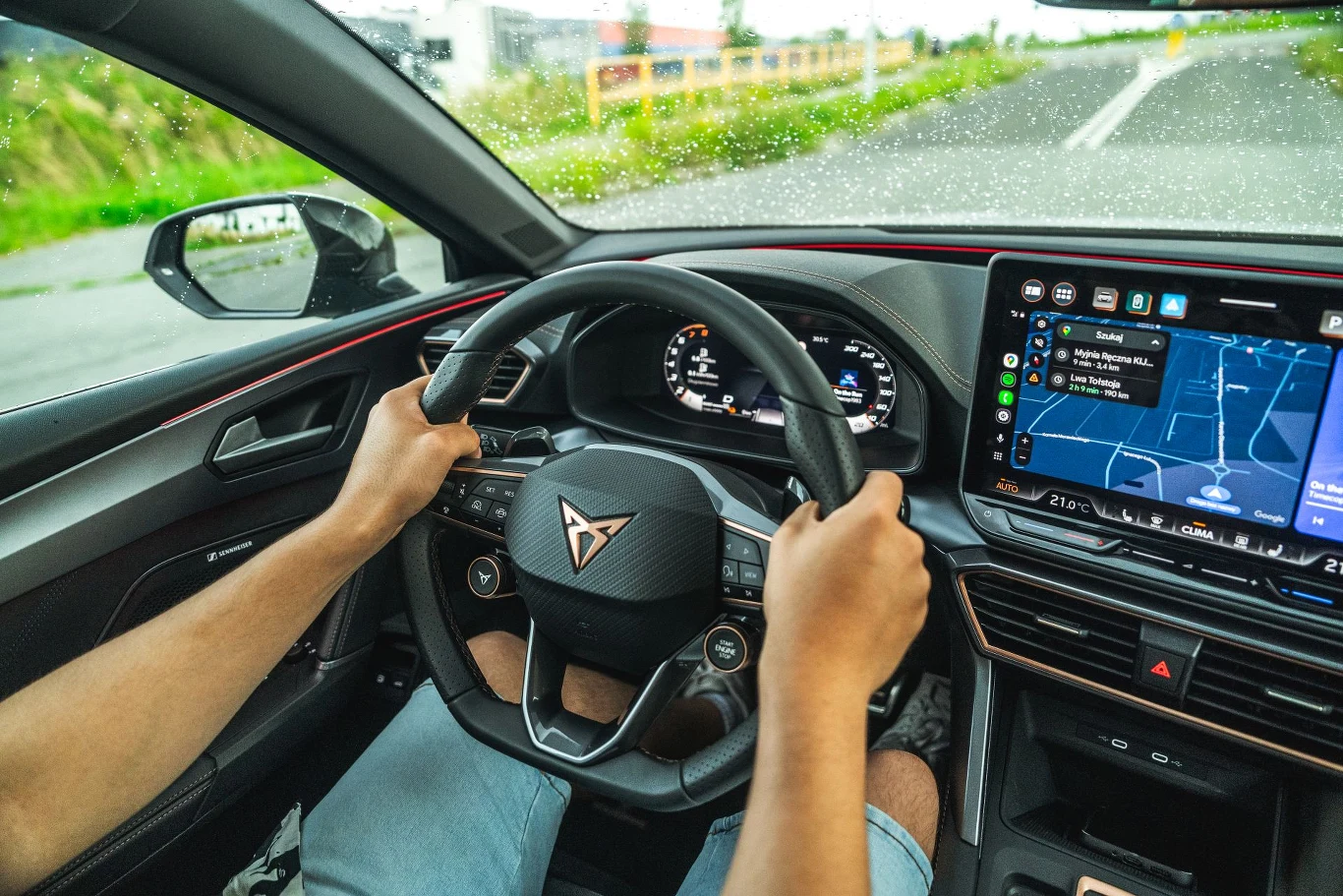 Odpowiednie obciążenie przedniej i tylnej osi idzie w parze z precyzyjnym układem kierowniczym, a także dodatkowym w tej wersji wyposażenia, adaptacyjnym zawieszeniem. Dzięki temu 204-konna Cupra Leon z hybrydą plug-in może dawać przyjemność z jazdy. Wnętrze nowoczesnego samochodu podczas jazdy, męskie ręce trzymają kierownicę z logo marki Cupra, na ekranie centralnym widoczny system nawigacji, za oknem fragment drogi i zieleni.