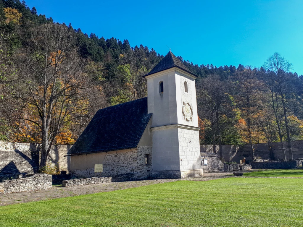 Czerwony Klasztor to urokliwe miejsce, które przenosi w czasy średniowiecza Stary, kamienny kościół z białą wieżą, położony na tle lasu i otoczony kamiennym murem, z zadbanym trawnikiem na pierwszym planie, w słoneczny dzień.