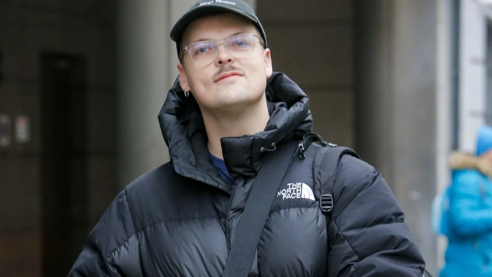 Błażej Król Mężczyzna w czarnej kurtce puchowej z logo The North Face, czapce z daszkiem oraz okularach, patrzy w obiektyw, mając za sobą rozmazane miejskie tło.