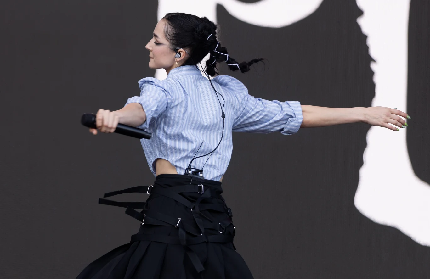 Poppy nagrała swoją wersję "Last Christmas" grupy Wham! Kobieta w stylowej, niebieskiej koszuli i czarnej spódnicy występuje na scenie, trzyma mikrofon, z rozpuszczonymi włosami splecionymi w ozdobne warkocze, rozkładając ramiona w dynamicznym geście.