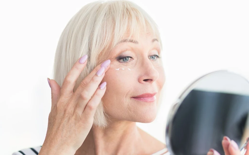 Pod oczy stosuj krem dedykowany tej partii ciała. Skład kremów jest delikatniejszy Kobieta o siwych włosach uśmiecha się lekko, stojąc przed lustrem i delikatnie nakłada krem pod oczy palcem, na jasnym tle.