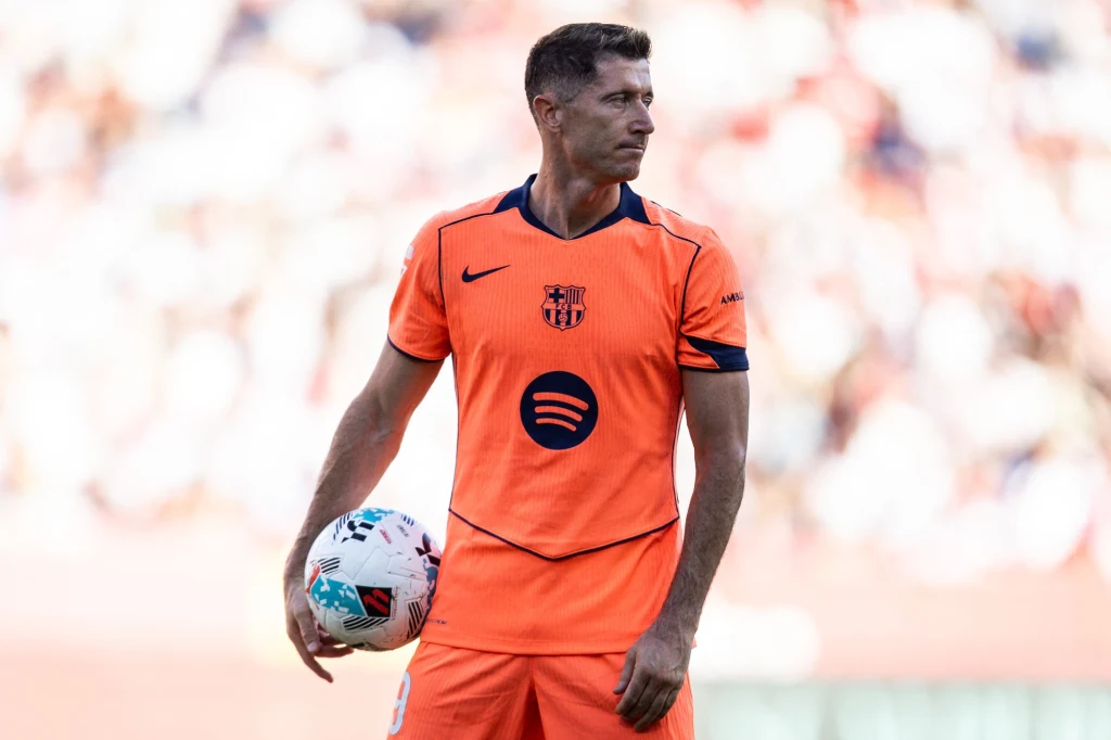 Piłkarz w pomarańczowym stroju FC Barcelony trzyma piłkę meczową, stoi na tle rozmytego stadionu podczas spotkania piłkarskiego.