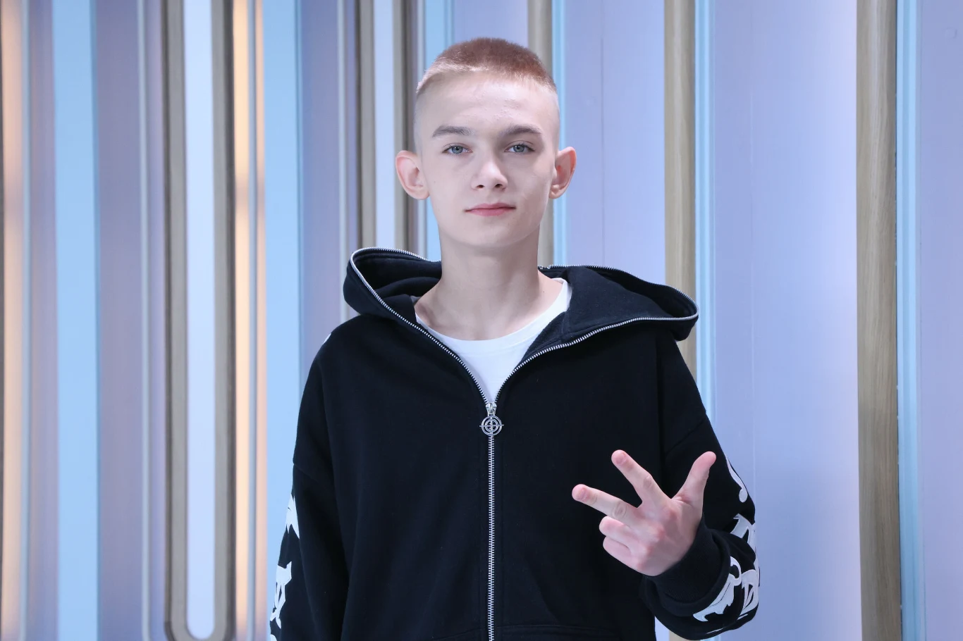 Bartek "Wasyl" Wasilewski wygrał program "Mam talent" w 2024 r. Młody mężczyzna z krótkimi włosami ubrany w czarną bluzę z białym zamkiem, pokazuje ręką znak pokoju, stoi na tle ściany z jasnymi pionowymi pasami.