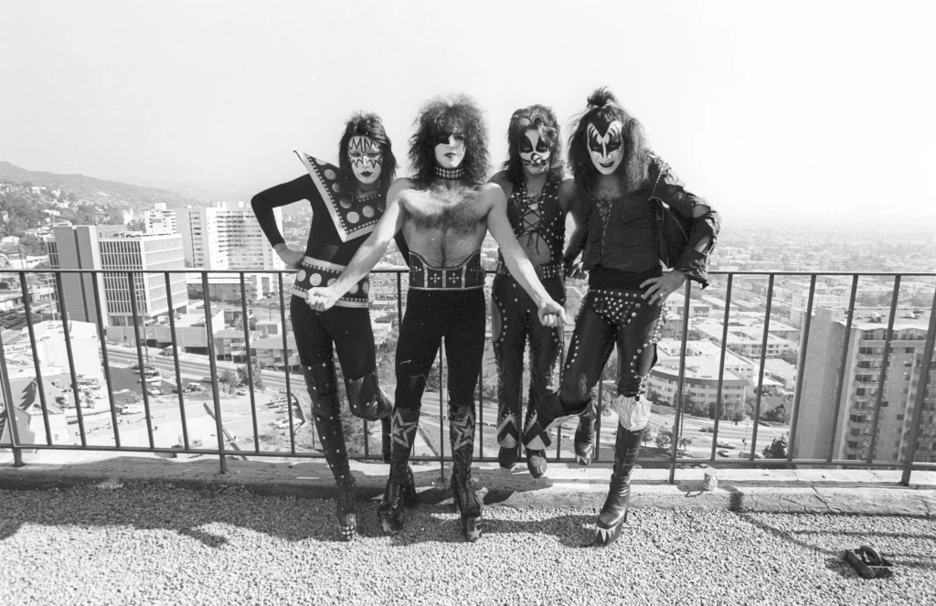 Czwórka mężczyzn ubranych w wyszukane stroje sceniczne z makijażem na twarzach pozuje na tle panoramy miejskiej, stojąc obok siebie na tarasie widokowym.