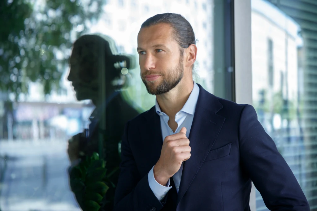 Grzegorz Krychowiak Elegancko ubrany mężczyzna w garniturze stoi przy oknie, z pewnym siebie wyrazem twarzy, patrząc w dal, a jego odbicie widoczne jest w szybie.