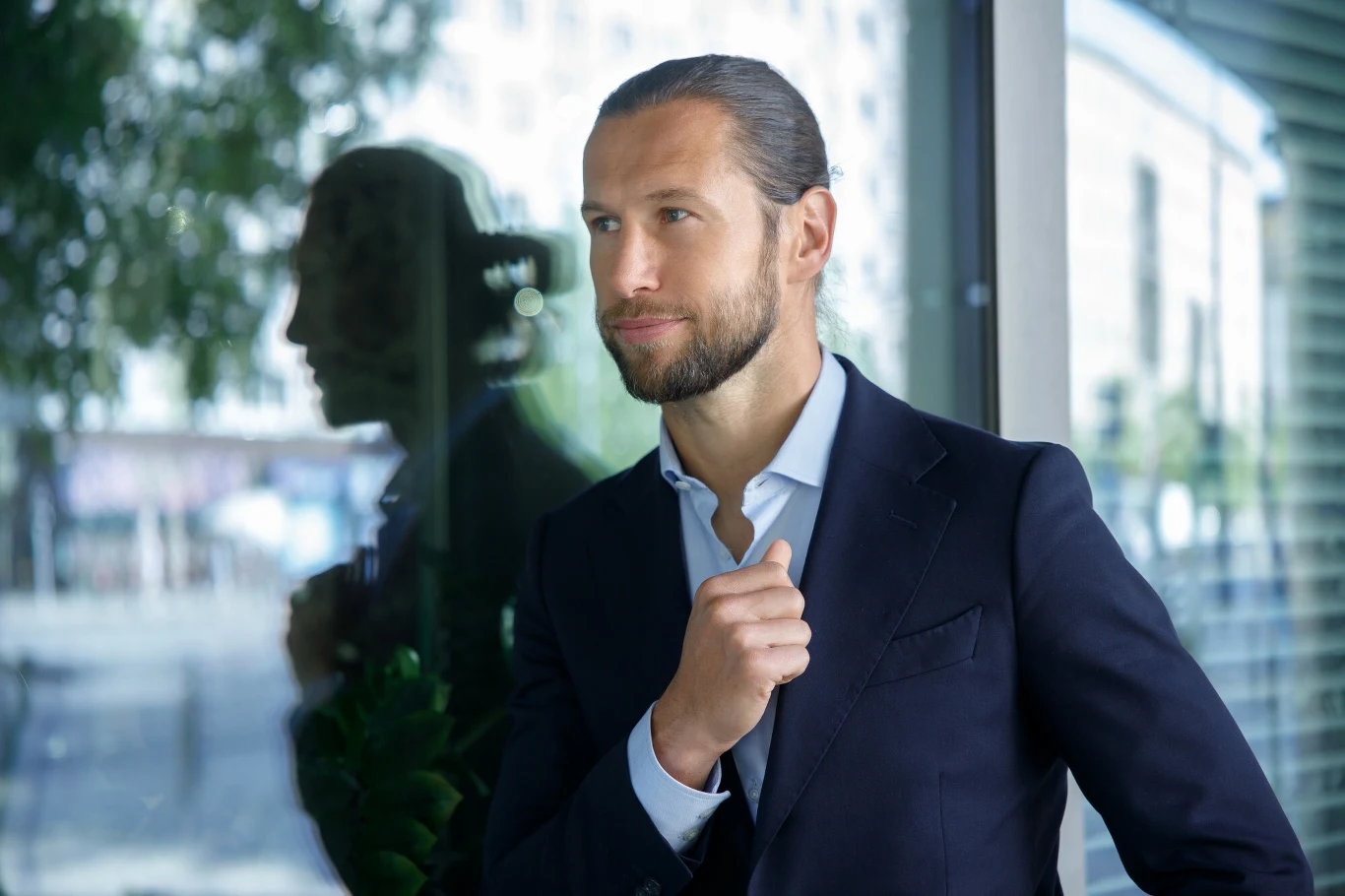 Elegancko ubrany mężczyzna w garniturze stoi przy oknie, z pewnym siebie wyrazem twarzy, patrząc w dal, a jego odbicie widoczne jest w szybie.