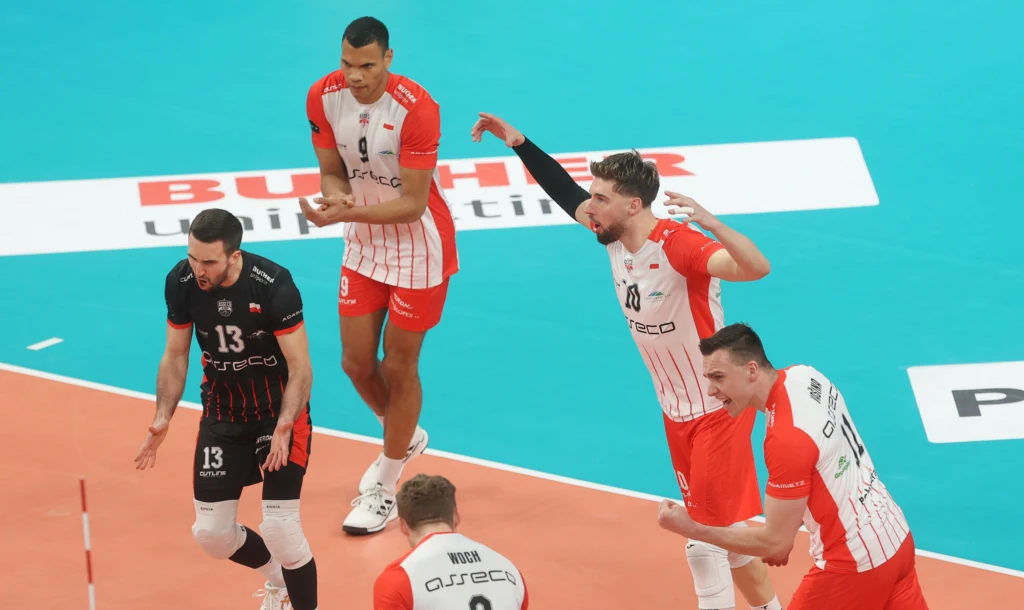 Grupa siatkarzy w dynamicznej akcji, zawodnicy wyrażają emocje po udanej akcji podczas meczu siatkówki, na kolorowym parkiecie z widocznymi oznaczeniami boiska.