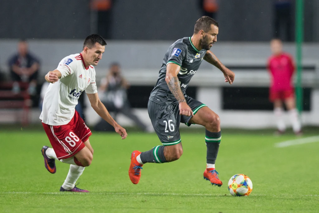 Błażej Augustyn przy piłce Dwóch piłkarzy w akcji podczas meczu piłki nożnej, jeden zawodnik w białej koszulce próbuje odebrać piłkę drugiemu graczowi w ciemnym stroju, którzy koncentrują się na rozgrywaniu piłki na boisku.