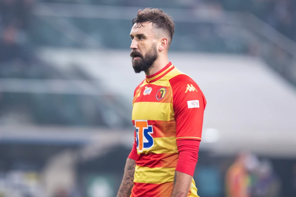 Błażej Augustyn Piłkarz w żółto-czerwonym stroju z logo sponsorów i klubowym herbem, stojący na boisku podczas meczu piłki nożnej, w tle rozmyte trybuny stadionu.