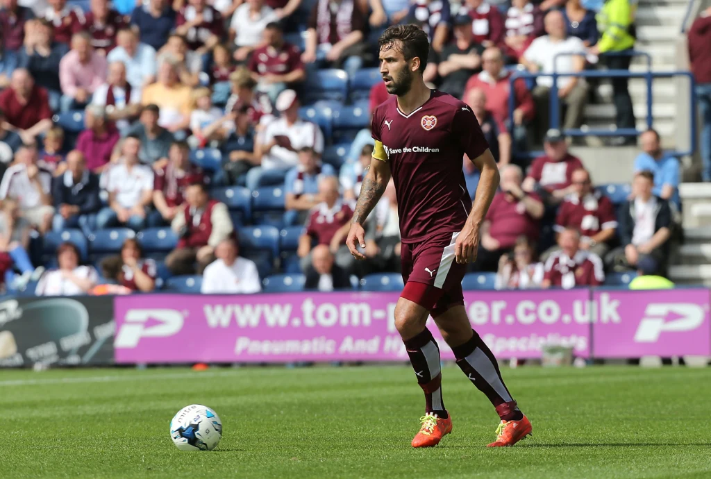 Błażej Augustyn Piłkarz w bordowym stroju z piłką przy nodze podczas meczu, w tle trybuny z kibicami.