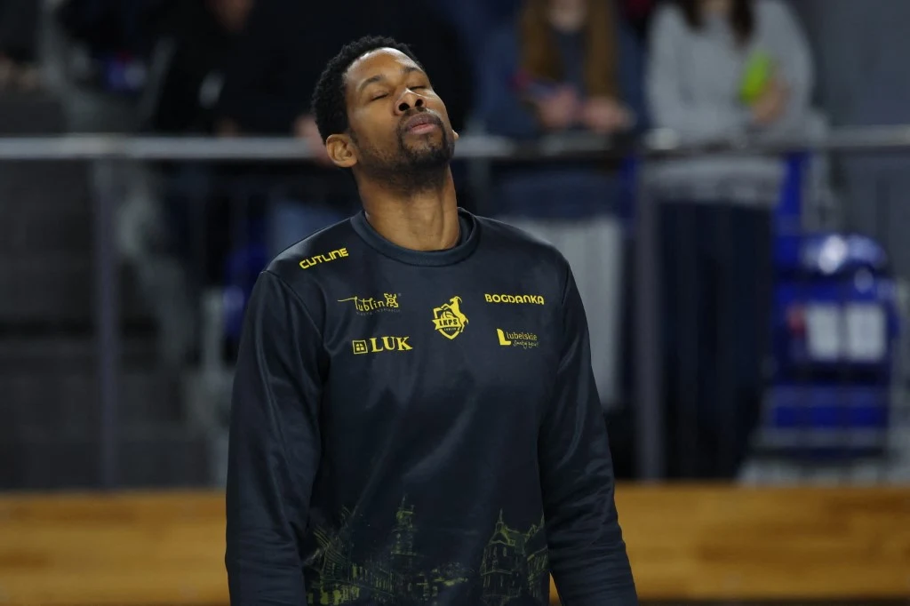 Wilfredo Leon Koszykarz w czarnej koszulce z zamkniętymi oczami, stojący na hali sportowej, w tle widownia i elementy infrastruktury sportowej.