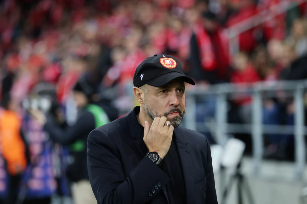 Mężczyzna z zarostem, ubrany w ciemną marynarkę i czapkę z daszkiem z klubowym emblematem, stoi na tle trybun stadionu wypełnionych publicznością ubraną na czerwono.