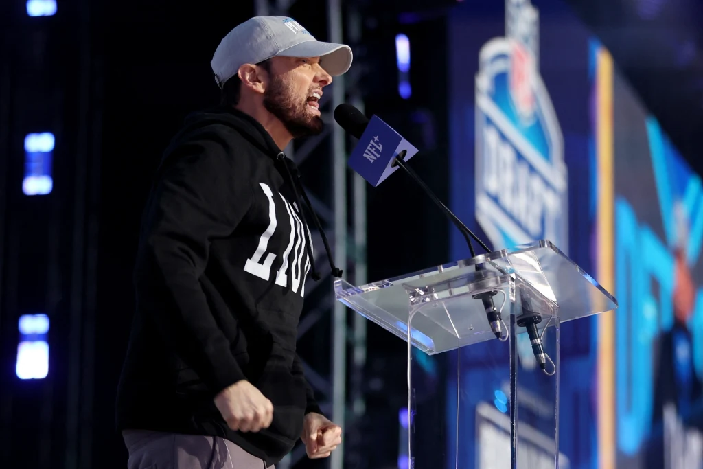 Eminem Mężczyzna w czarnej bluzie i jasnej czapce z daszkiem przemawia przy przezroczystej mównicy na tle logo eventu sportowego, scena oświetlona niebieskim światłem.