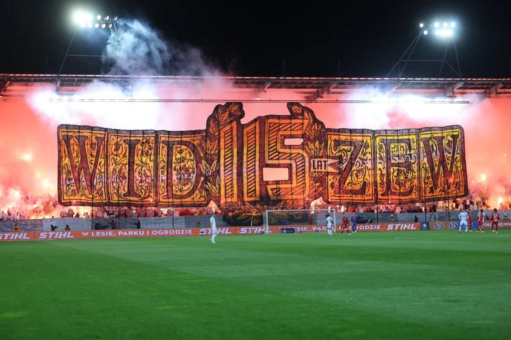 Transparent o treści 'Widzew 115 lat' rozciągnięty na trybunach stadionu piłkarskiego, podświetlony racami, z piłkarzami na murawie oraz tłumem kibiców świętujących rocznicę klubu.