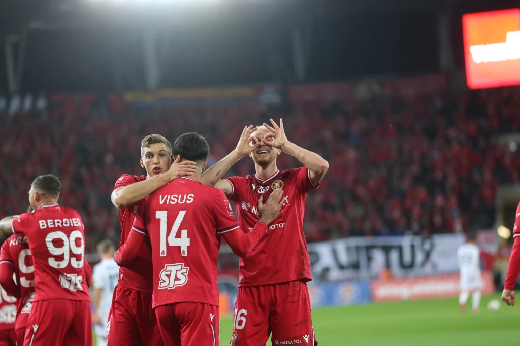 Piłkarze drużyny w czerwonych strojach świętują zdobycie bramki na tle trybun z kibicami, jeden z zawodników wykonuje gest rękami, pozostali obejmują się i wyrażają radość.