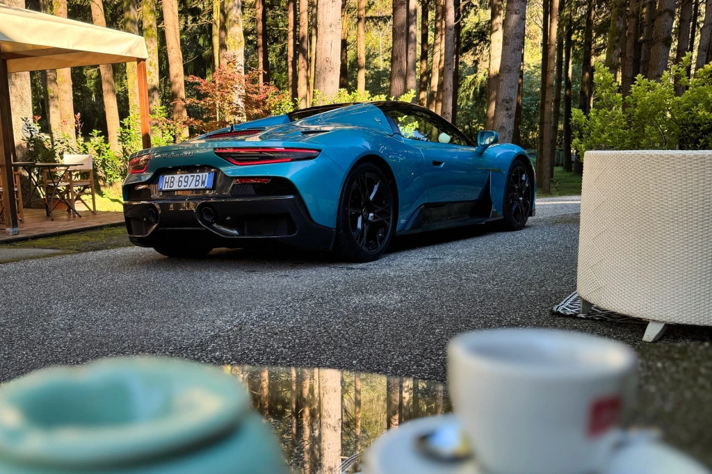 Poranek w Forte dei Marmi. Kawa jeszcze gorąca, a 630-konne Maserati już gotowe do drogi Turkusowy samochód sportowy na tle wysokich drzew w lesie, zaparkowany obok stolika kawiarnianego z filiżankami na pierwszym planie. Jasne światło wydobywa intensywny kolor auta i tworzy kontrast z zielenią roślin.