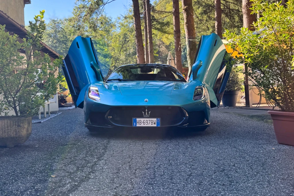 Drzwi unoszą się do góry jak skrzydła. Włoski teatr zaczyna się jeszcze zanim silnik Nettuno ożyje Luksusowe sportowe auto o niebieskim kolorze z otwartymi ku górze drzwiami, ustawione na podjeździe w otoczeniu zieleni i wysokich drzew, po bokach donice z roślinami, rejestracja włoska.