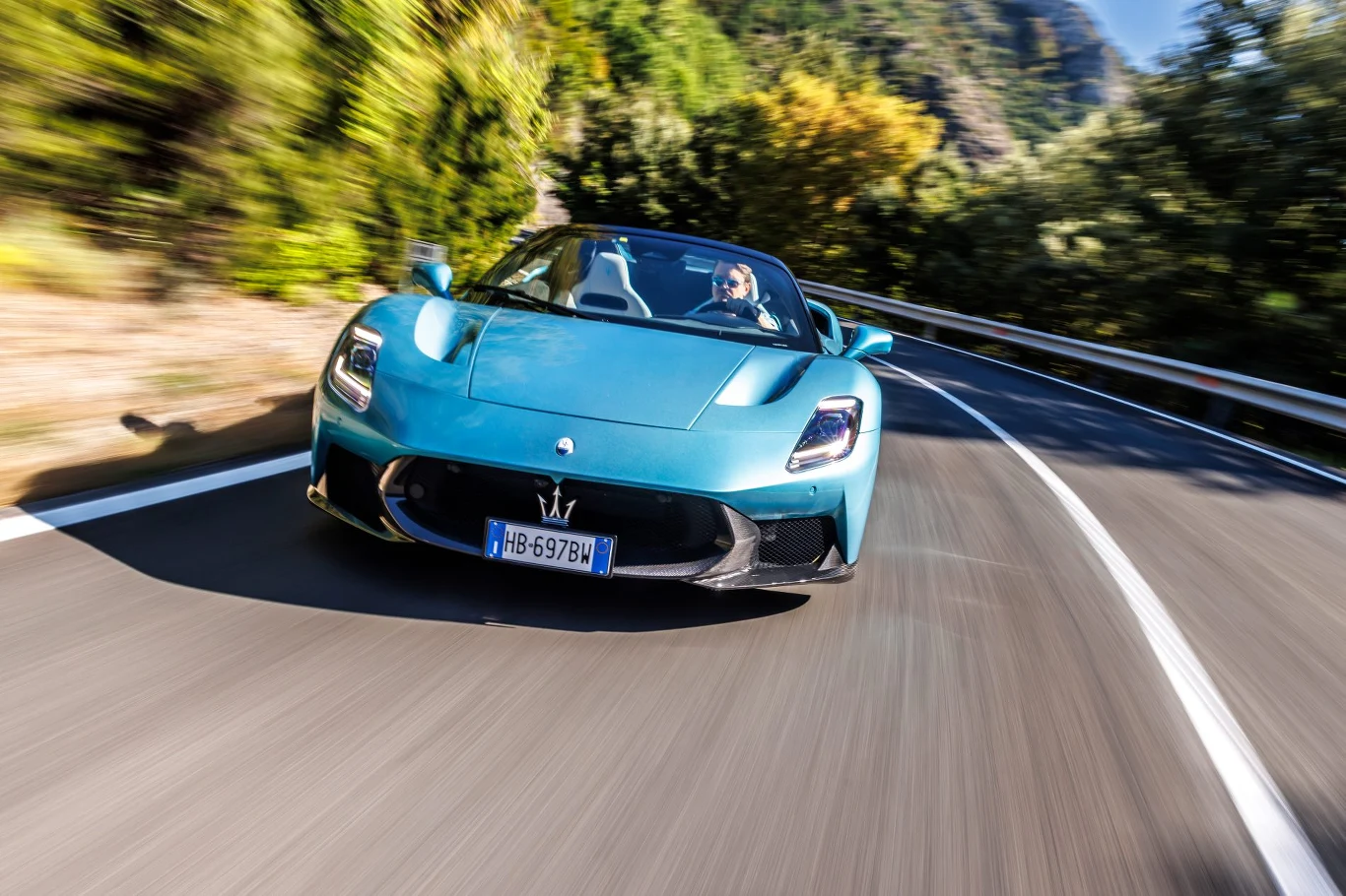 Sportowy niebieski samochód Maserati MC20 jadący dynamicznie po krętej górskiej drodze otoczonej zielenią, w słonecznej scenerii z widocznymi rozmazanymi liniami ruchu podkreślającymi prędkość auta.