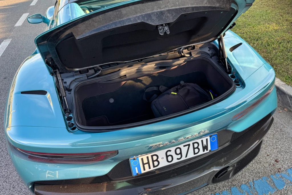 Z tyłu MCPura Cielo mieści się około 100 litrów bagażu. W praktyce – jedna miękka torba i tyle Otwarty tylny bagażnik sportowego samochodu w kolorze turkusowym, w środku bagażu znajduje się czarna torba podróżna, widoczna tablica rejestracyjna oraz fragment asfaltu i trawnika.