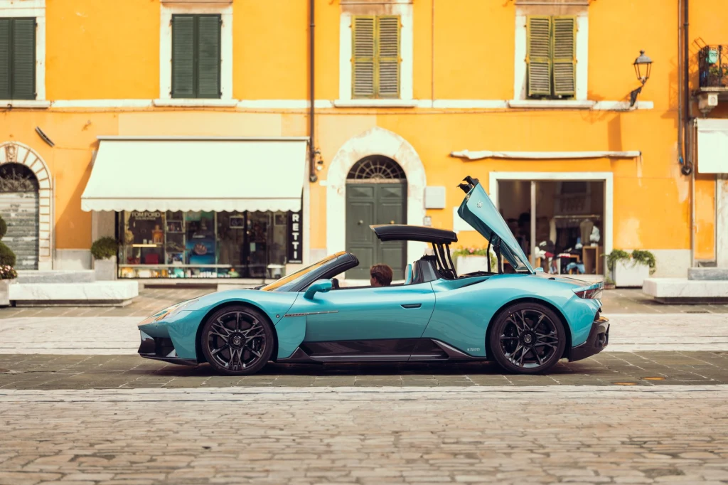 Dach znika za plecami kierowcy w 12 sekund Jasnoniebieski luksusowy samochód sportowy z otwartym dachem, parkujący na brukowanym placu miejskim, na tle żółtej kamienicy z białymi oknami i roletami, kierowca siedzi w środku.