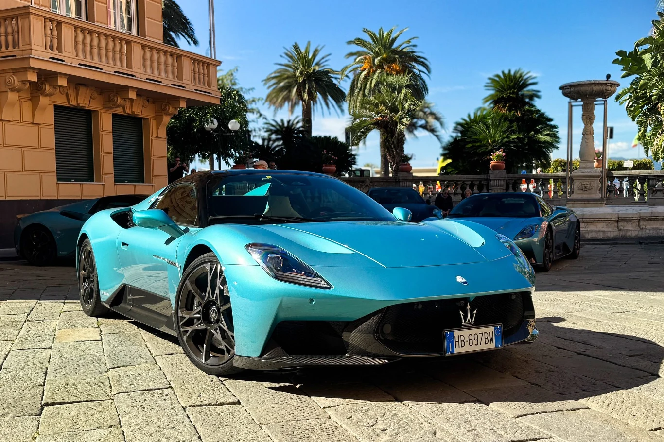 Turkusowy sportowy samochód Maserati MC20 zaparkowany na brukowanym placu przy eleganckim budynku z palmami i fontanną w tle, po lewej stronie widoczna część drugiego pojazdu.
