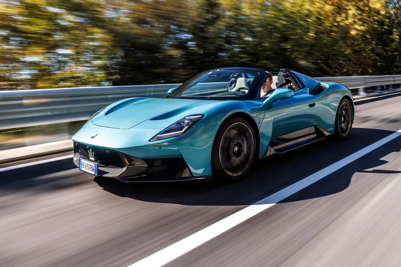 Sportowy samochód Maserati MC20 z otwartym dachem sunie szybko po asfaltowej drodze, otoczony rozmytym tłem drzew, podkreślającym prędkość i dynamiczny charakter auta.