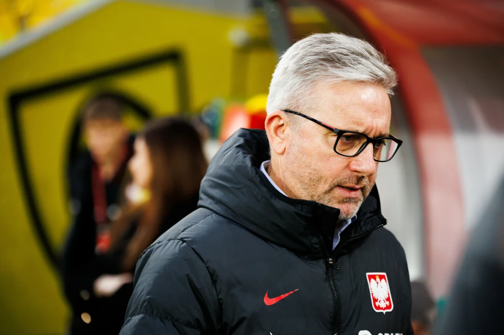 Mężczyzna w okularach i kurtce z herbem Polski, stojący w sportowym otoczeniu, w tle rozmyte sylwetki osób oraz fragment ławki rezerwowych.