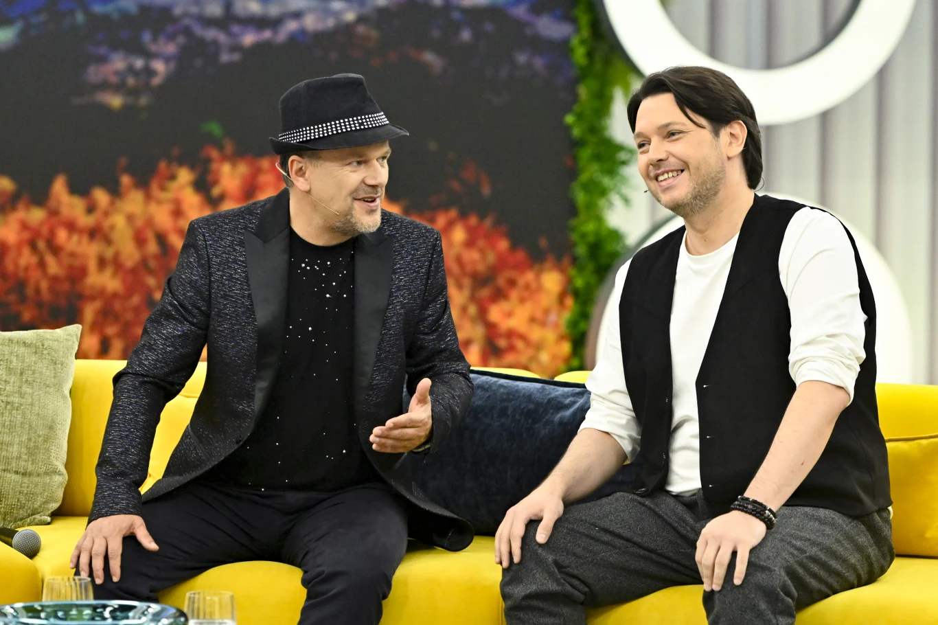 Krzysztof "K.A.S.A." Kasowski i Gabriel Fleszar w programie "halo tu polsat" Dwóch mężczyzn siedzi na żółtej sofie i rozmawia ze sobą z uśmiechami na twarzach w jasnym, nowoczesnym wnętrzu z dekoracjami i roślinnością w tle.