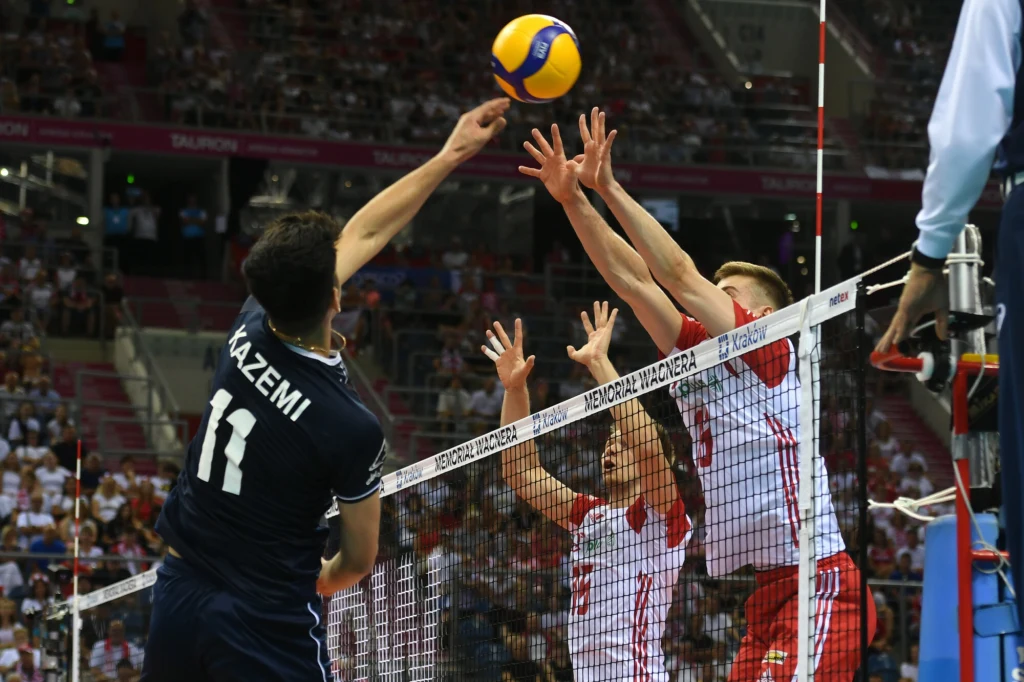 Zawodnik drużyny w granatowym stroju atakuje piłkę nad siatką, podczas gdy dwóch zawodników w biało-czerwonych strojach blokuje po drugiej stronie. W tle widoczna publiczność na trybunach.