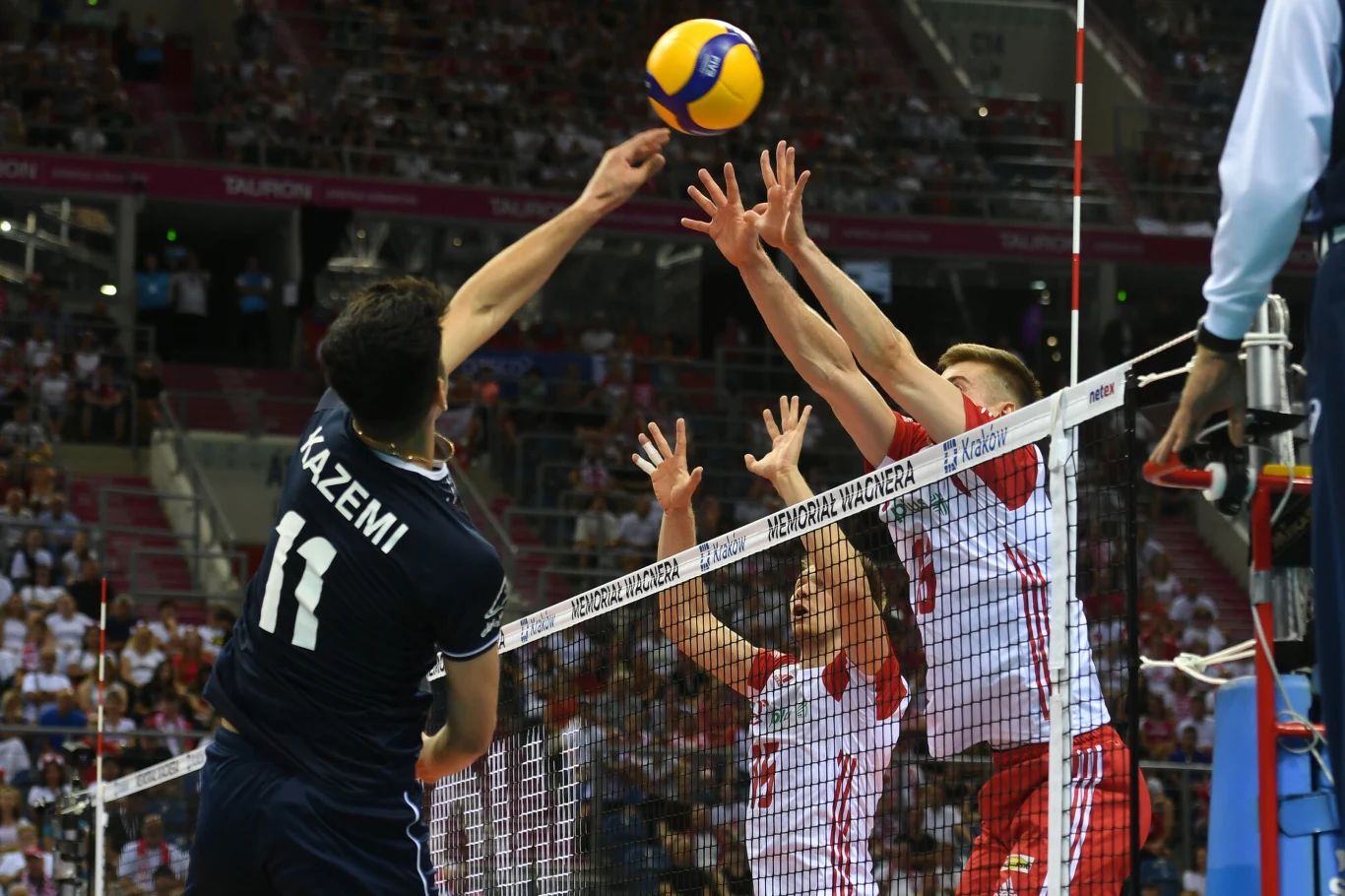 Zawodnik drużyny w granatowym stroju atakuje piłkę nad siatką, podczas gdy dwóch zawodników w biało-czerwonych strojach blokuje po drugiej stronie. W tle widoczna publiczność na trybunach.