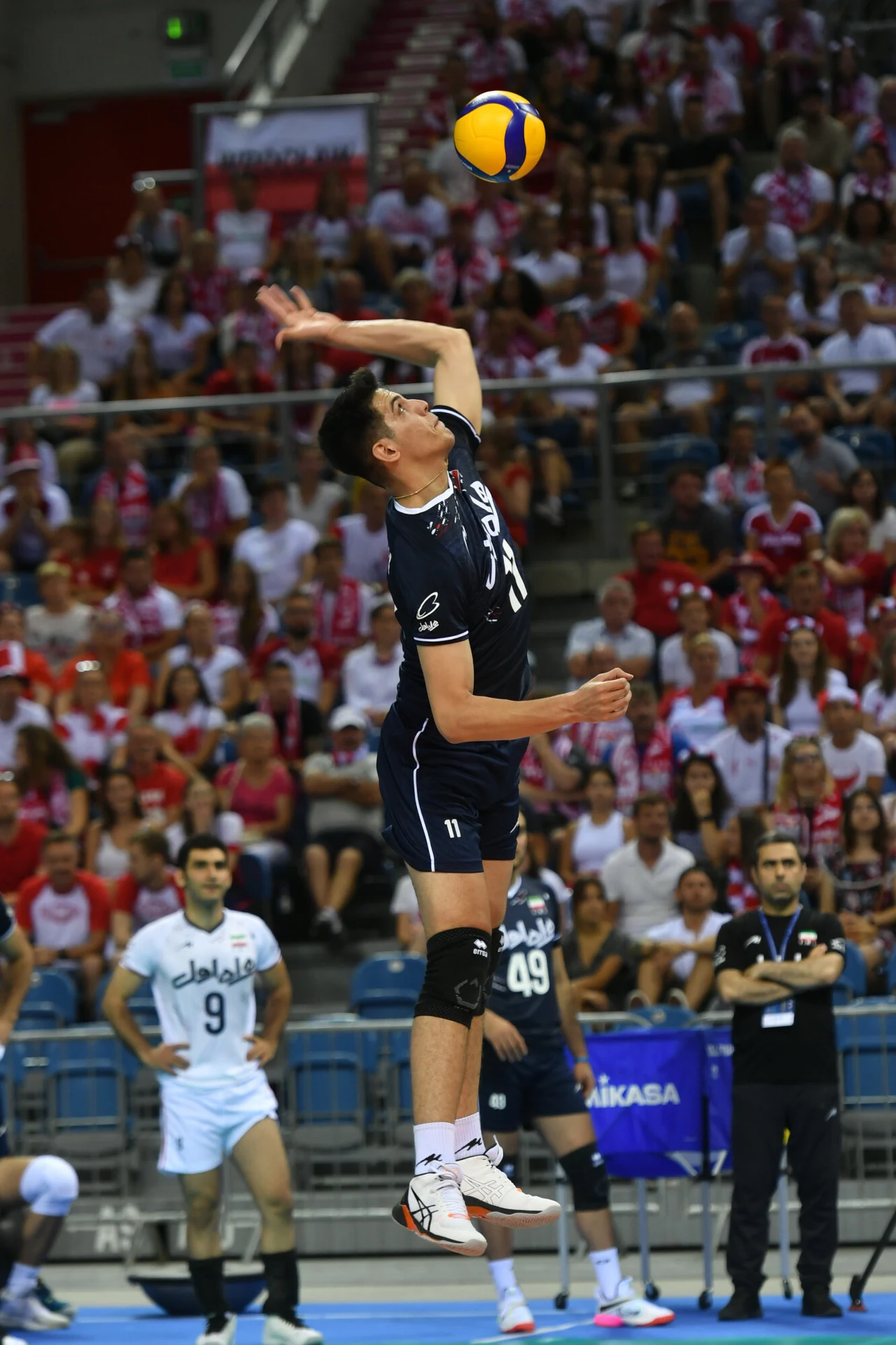 Saber Kazemi Zawodnik w ciemnym stroju wykonuje serwis podczas meczu siatkówki, dynamiczna sylwetka w locie, w tle widoczna pełna widownia oraz inni siatkarze i trenerzy.