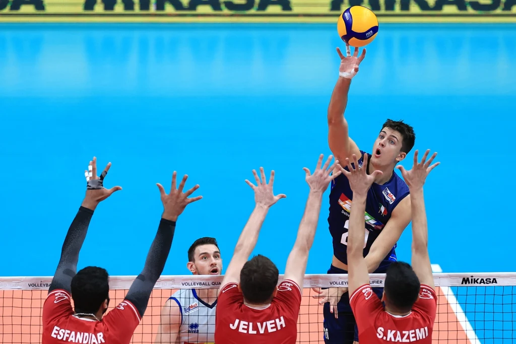 Saber Kazemi Zawodnik drużyny siatkarskiej wykonuje atak ponad siatką, próbując obić blok trzech rywali w czerwonych koszulkach na tle niebieskiego boiska.