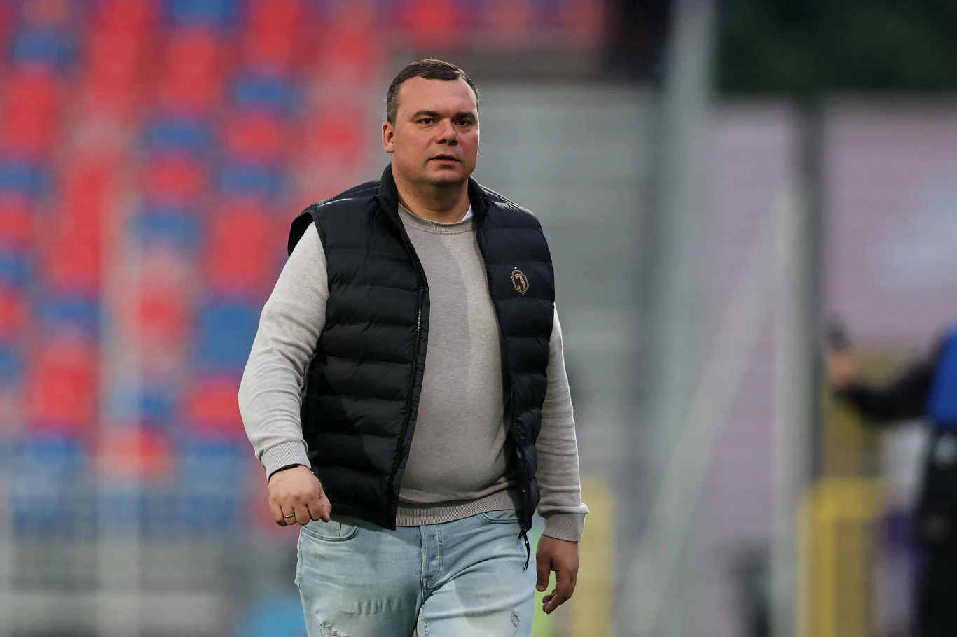 Mężczyzna w czarnej kamizelce i jasnych dżinsach idzie po stadionie, w tle widoczne puste krzesełka w kolorach czerwonym i niebieskim.