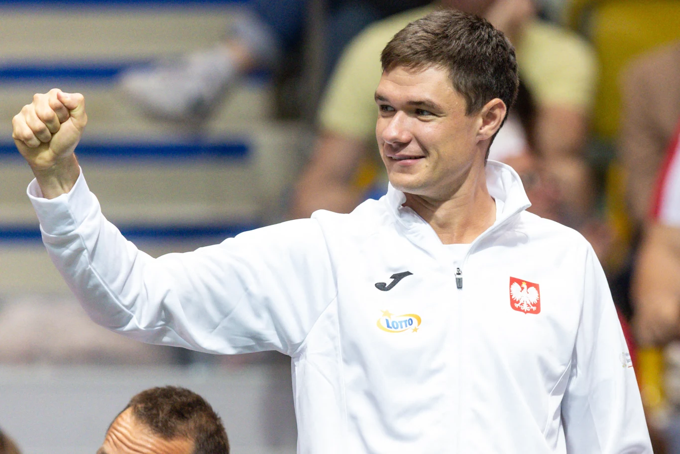 Kamil Majchrzak Mężczyzna ubrany w biały dres z polskim godłem i logo sponsora unosi zaciśniętą pięść w geście triumfu lub pozdrowienia, w tle widoczne rozmyte sylwetki ludzi i żółte trybuny.