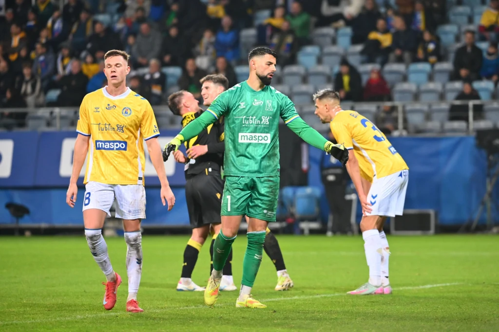 Piłkarze w żółtych koszulkach oraz bramkarz w zielonym stroju stoją na boisku podczas meczu, część zawodników wyraża emocje po rozegranej akcji, w tle widoczna publiczność na trybunach.