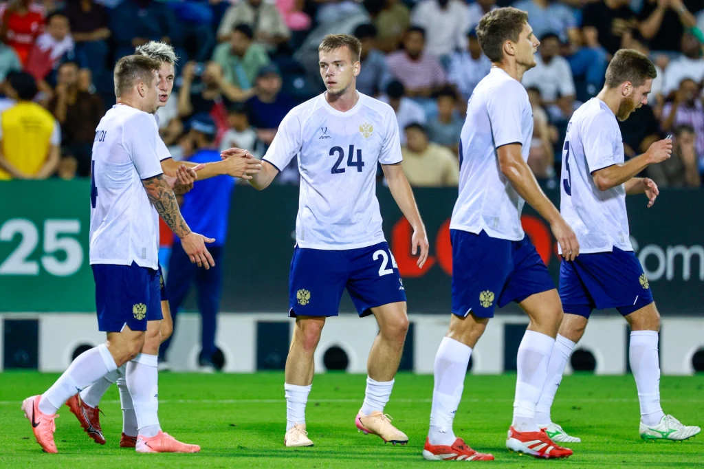 Grupa piłkarzy w białych koszulkach i niebieskich spodenkach celebruje akcję na boisku, jeden z zawodników podaje dłoń koledze z drużyny, na trybunach widoczna publiczność.