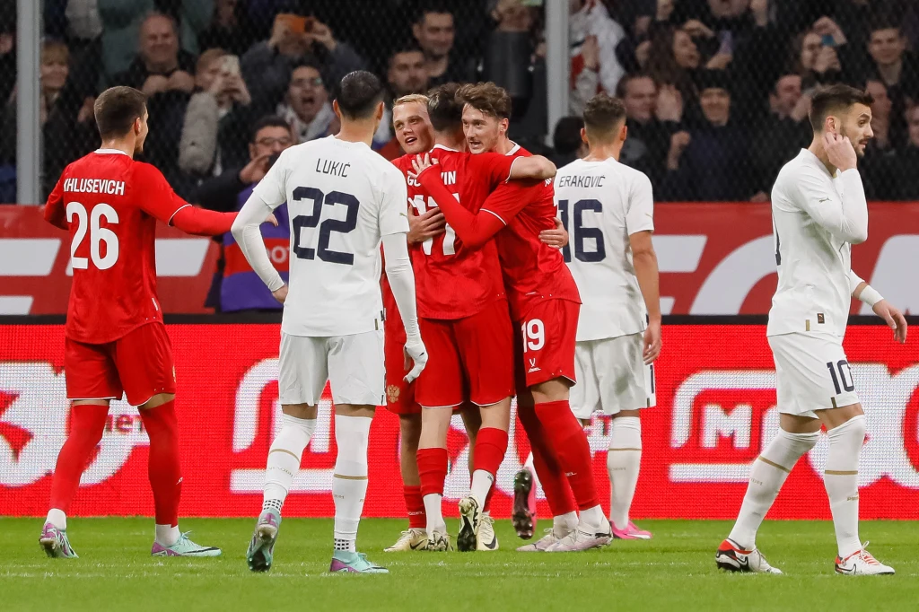 Grupa piłkarzy w czerwonych i białych strojach podczas meczu piłkarskiego, kilku zawodników w czerwonych koszulkach świętuje zdobycie gola i obejmuje się na środku boiska, wokół nich widać rywali i kibiców na trybunach.