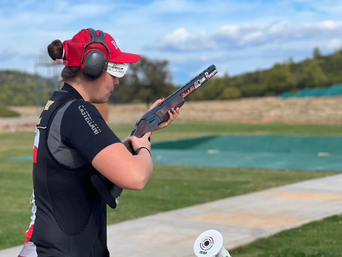 Kobieta w sportowej odzieży i nausznikach ochronnych celuje z dubeltówki na otwartym terenie strzelnicy, w tle widoczna zieleń i niebo z lekkim zachmurzeniem.